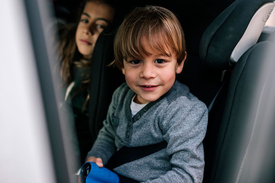A young boy smiles as he sits in the back seat of the car buckled into a car seat. His older sister sits next to him appearing behind the car seat.