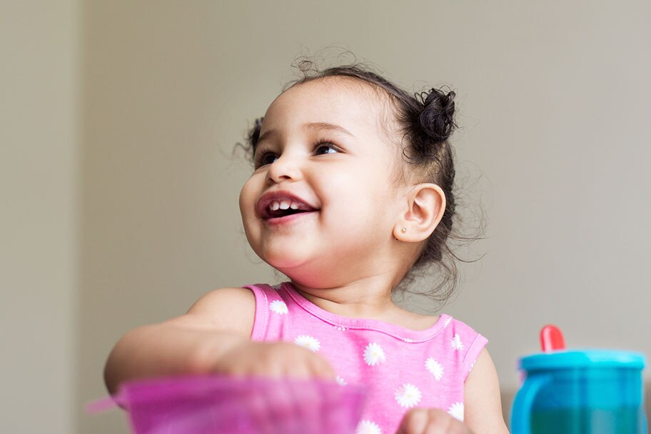 A 1 year old girl smiles with her hair in buns and a sippy cup beside her while she turns around in her chair.
