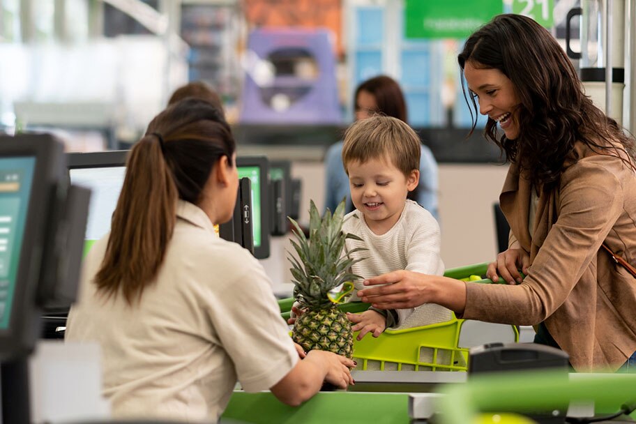 A young boy sitting in a grocery shopping cart helps his smiling mother pass a fresh pineapple to a cashier at the checkout.