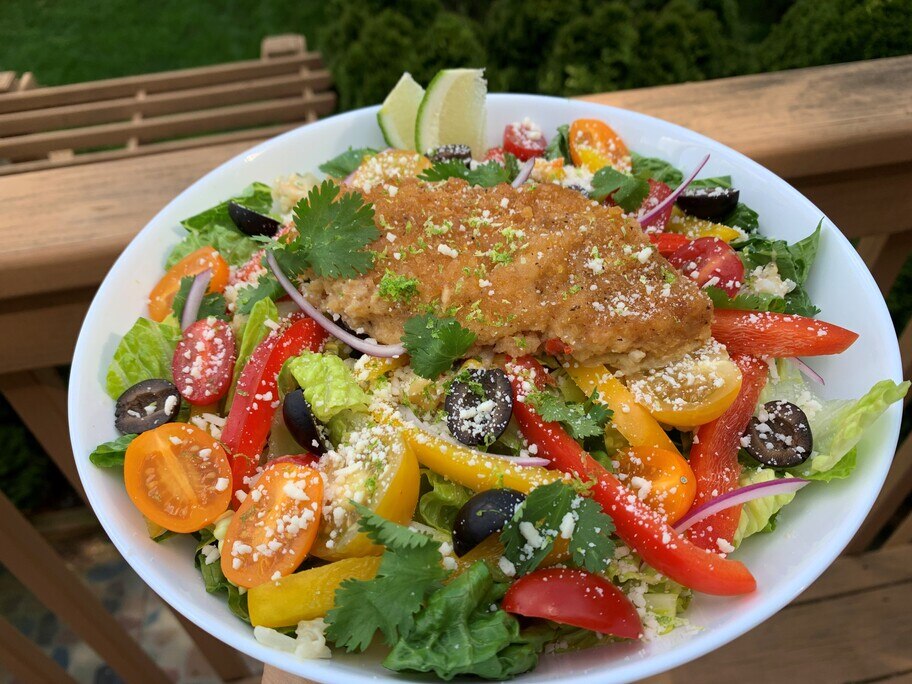 Un plato con una ensalada romana cubierta con pimientos, tomates, aceitunas, cebolla, queso y un filete de pescado con corteza de tortilla. 