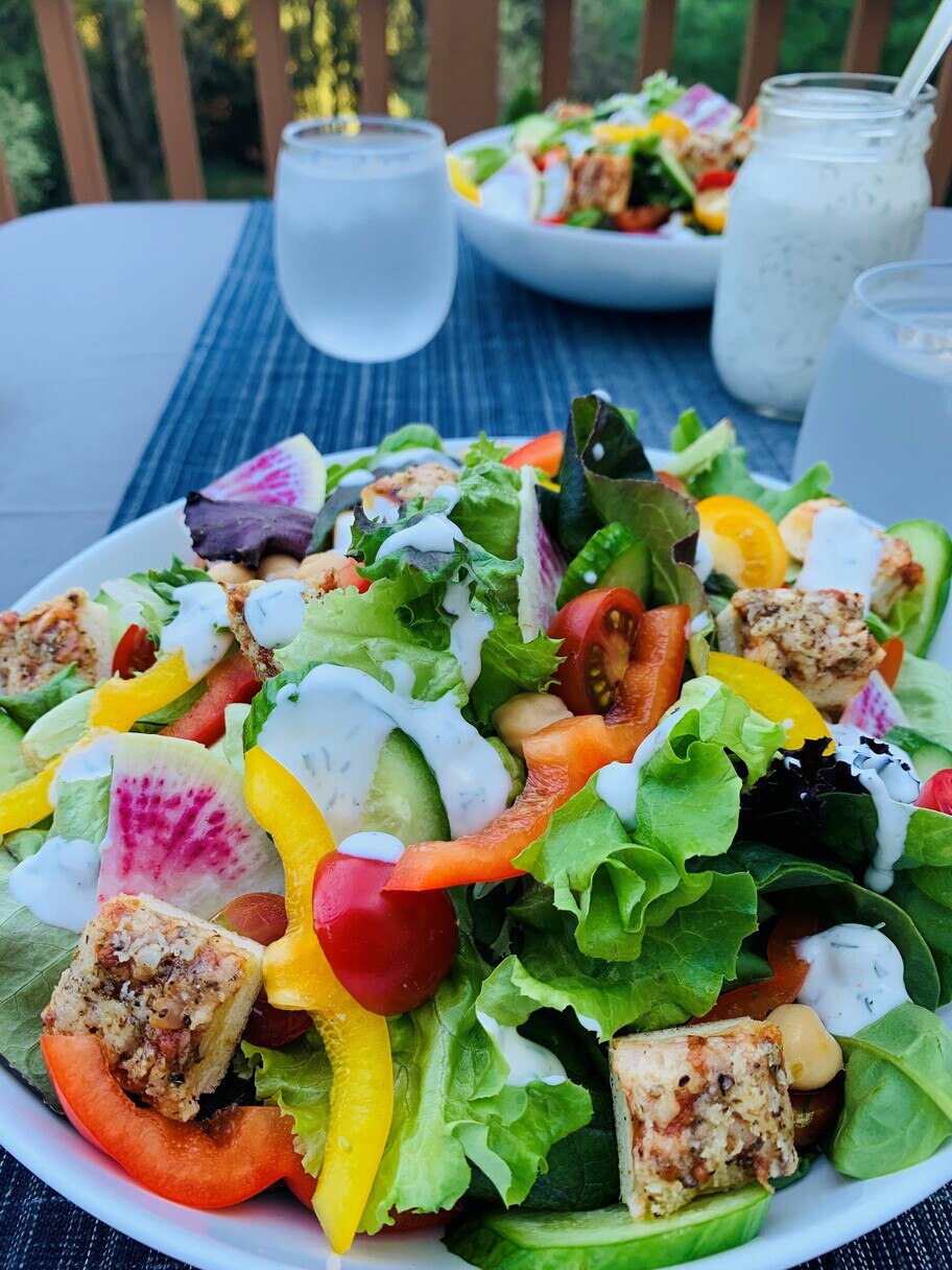Una mesa de patio con un frasco de aderezo, vasos de agua y platos de ensalada con morrones, tomates, pepinos y vegetales.