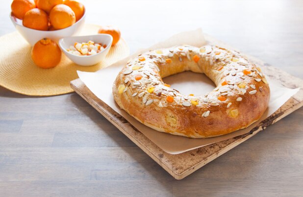 Una Rosca de Reyes sobre papel encerado junto a un tazón de naranjas y fruta deshidratada.