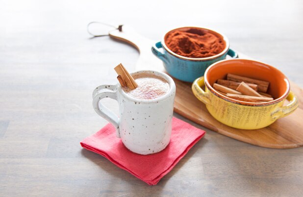 Una taza de chocolate caliente estilo mexicano junto a tazones de chile en polvo y ramitas de canela.