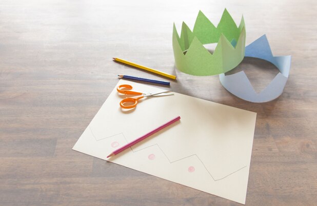 Dos coronas de papel junto a papel construcción, tijeras y lápices de colores.