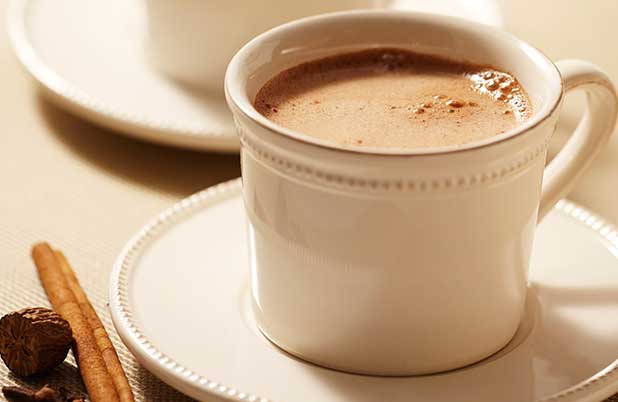 Una taza blanca de ABUELITA con un platillo blanco junto con ramitas de canela.