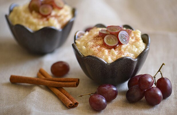  Tazones color negro con arroz con leche estilo chileno cubierto con uvas en rebanadas junto a ramitas de canela y uvas enteras.