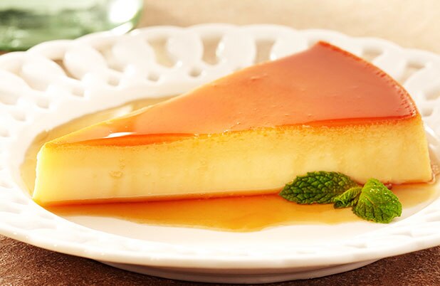 Una rebanada triangular de flan en un plato blanco con un fondo de caramelo y guarnición de menta.