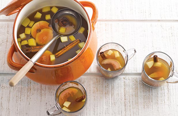 Una olla naranja de ponche navideño rodeada de tazas de vidrio con ponche y fruta.