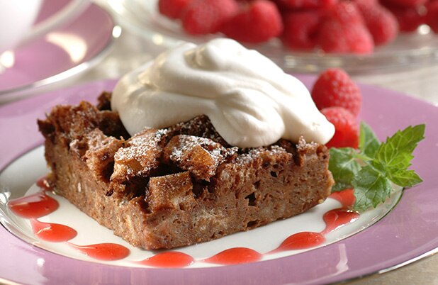 Una rebanada cuadrada de budín de pan moca en un plato con topping batido y frambuesas frescas.