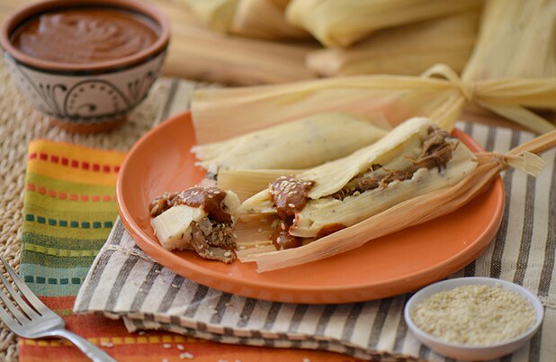 Un tamal de res abierto sobre un plato de cerámica junto a tazones de mole y condimentos.