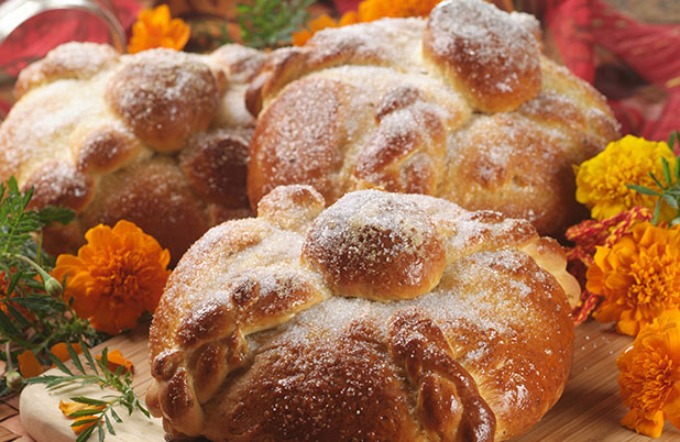 Tres hogazas de Pan de Muerto espolvoreadas con azúcar y rodeadas de flores.
