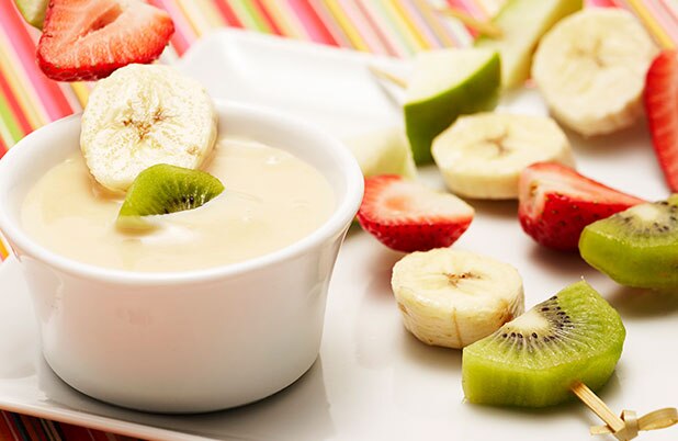 Brochetas de plátano, kiwi, fresa y manzana en un plato junto a una taza de salsa para comer con la fruta.