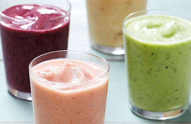 Smoothies de frutas colores púrpura, verde, rosado y amarillo en vasos sobre una mesa color azul.