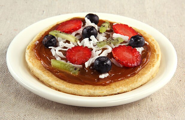 Una pizza de fruta hecha con un panqueque, chocolate untable, fruta fresca y coco rallado.