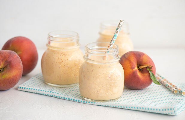 Tres frascos de vidrio llenos de batido de durazno junto a un durazno en una servilleta de tela.