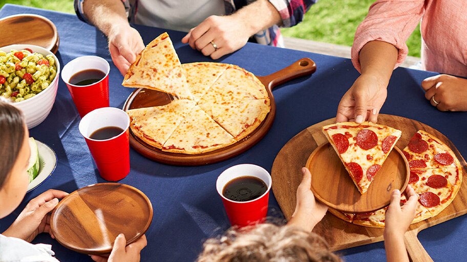 Una familia sentada en una mesa, con un tazón de pasta y platos de madera, reparte rebanadas de pizzas de queso y pepperoni.