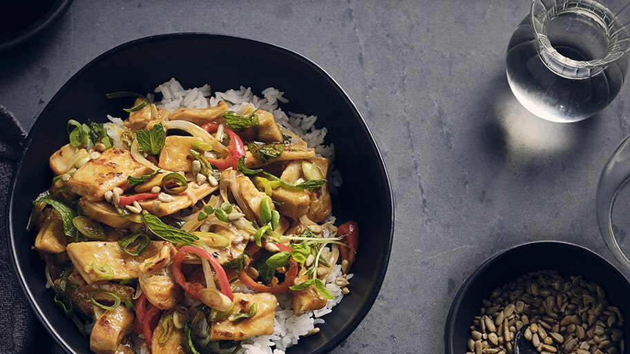 Un tazón de salteado de Mindful Chik'n con cebolletas y semillas de girasol cerca de un vaso de agua y un tazón de semillas.