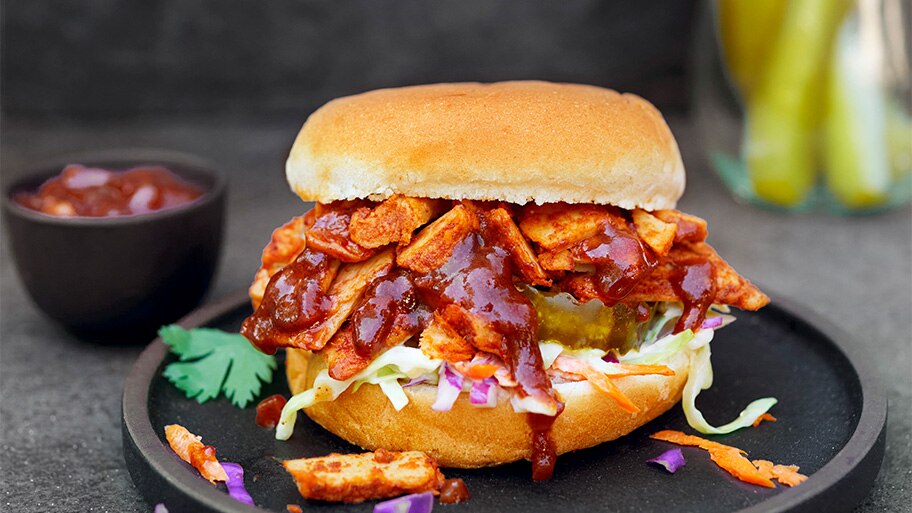 Un envase con pepinillos y uno de ketchup detrás de un slider de Chik'n con ensalada de repollo sobre un plato negro.