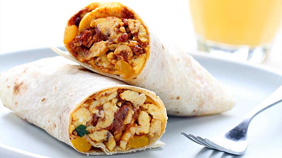 Burrito de desayuno cortado relleno de huevos, chorizo y verduras sobre un plato azul y un vaso de jugo de naranja al fondo.