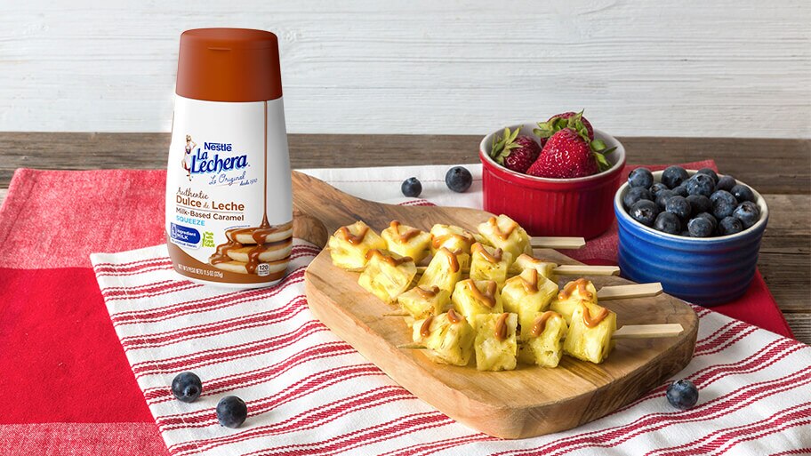 Brochetas de piña asada con dulce de leche por encima sobre una tabla con bayas y una botella de dulce de leche La Lechera.