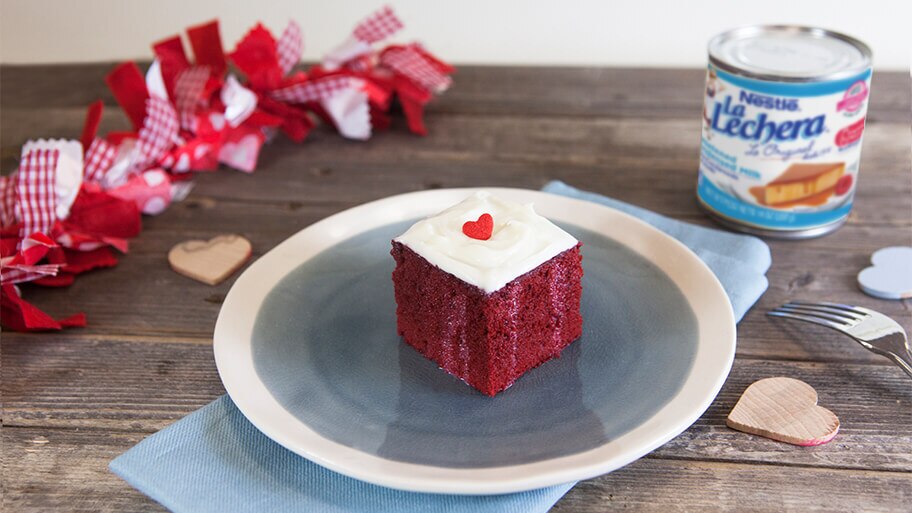 Pastel de terciopelo rojo bañado con queso crema decorado con un corazón rojo sobre una mesa con una lata de La Lechera.