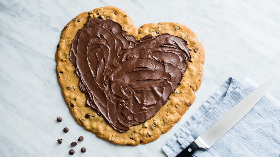 Pastel galleta en forma de corazón con glaseado de chocolate junto a servilleta rosa y cuchillo.