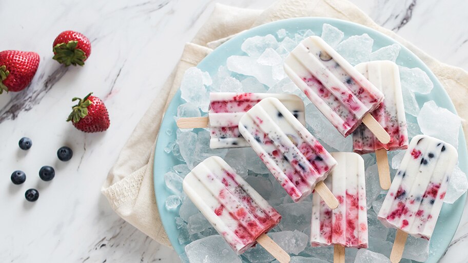 Berry popsicles on a bowl of ice near scattered blueberries and strawberries on a marble countertop.