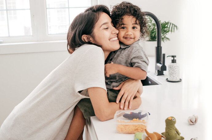 A young curly haired chil sits on the kitchen counter beside toys and snacks smiling while his young mom smiles and hugs him.