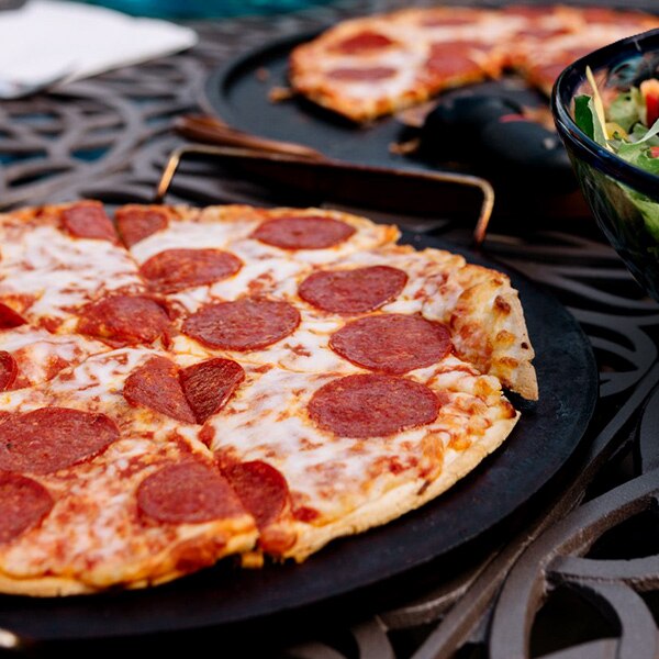 Original Thin Crust pepperoni pizza is cooling on a cast iron baking sheet on an oven beside another pizza and side salad.