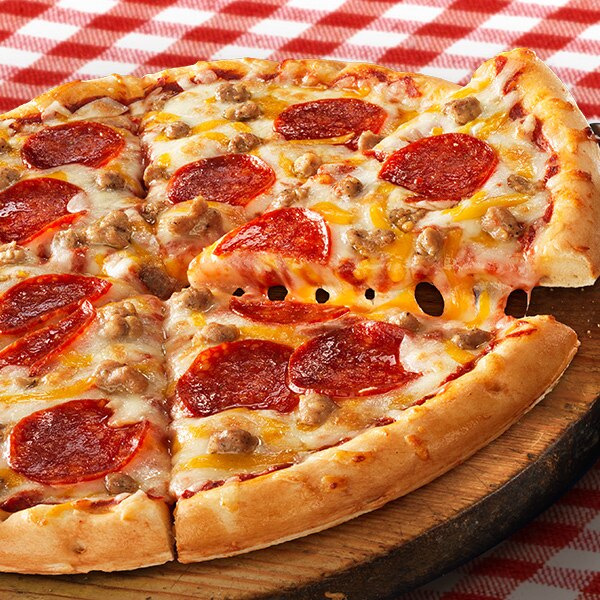 A slice of three meat Rising Crust pizza lifts from the pizza on a round wooden serving tray and classic red tablecloth.