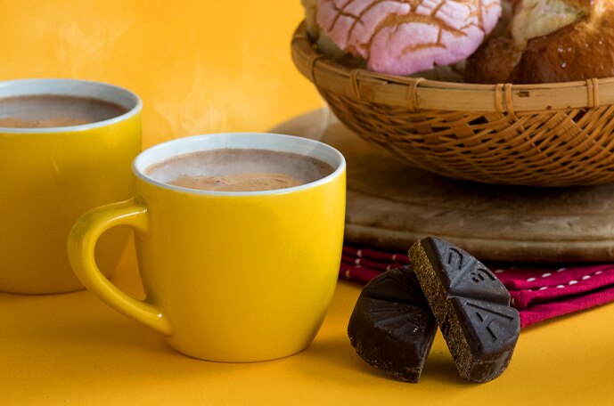 Two yellow mugs of hot chocolate beside a basket of conchas and ABUELITA tablets.