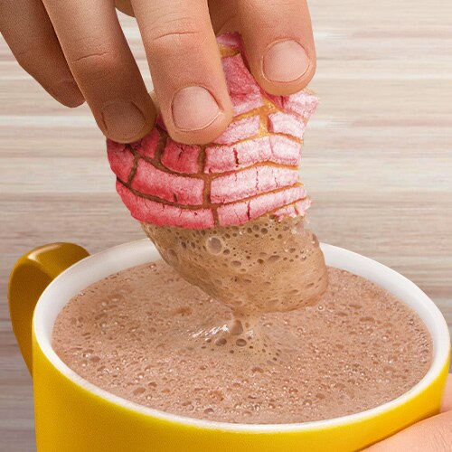 A hand dips a pink concha into a yellow mug with chocolate. 