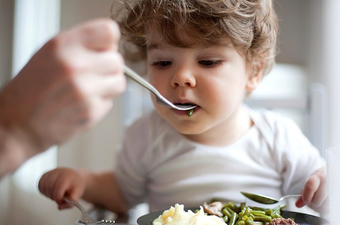 A hand spoon feeds a young boy mashed potatoes and green beans.