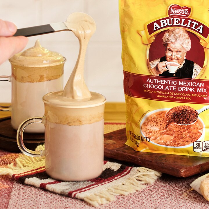 A spoon tops off a dalgona coffee on an Aztec inspired coaster beside a package of ABUELITA drink mix on a cutting board.