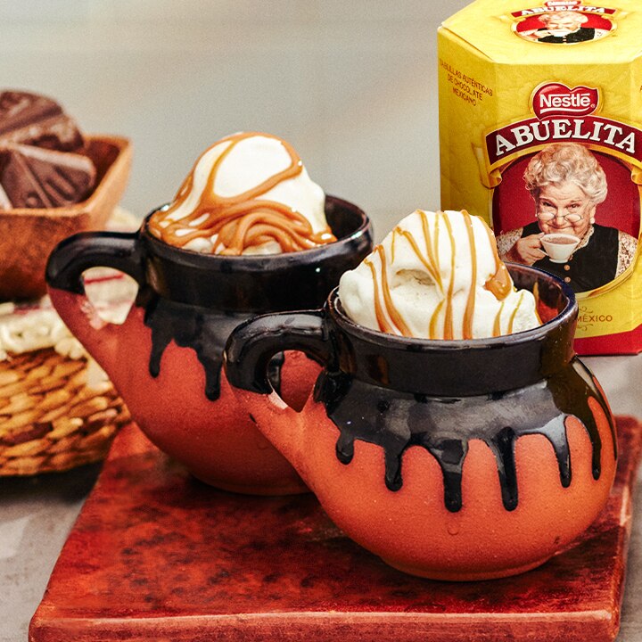 Two ceramic mugs filled with ice cream and caramel surrounded by cinnamon sticks with ABUELITA tablets and packaging.