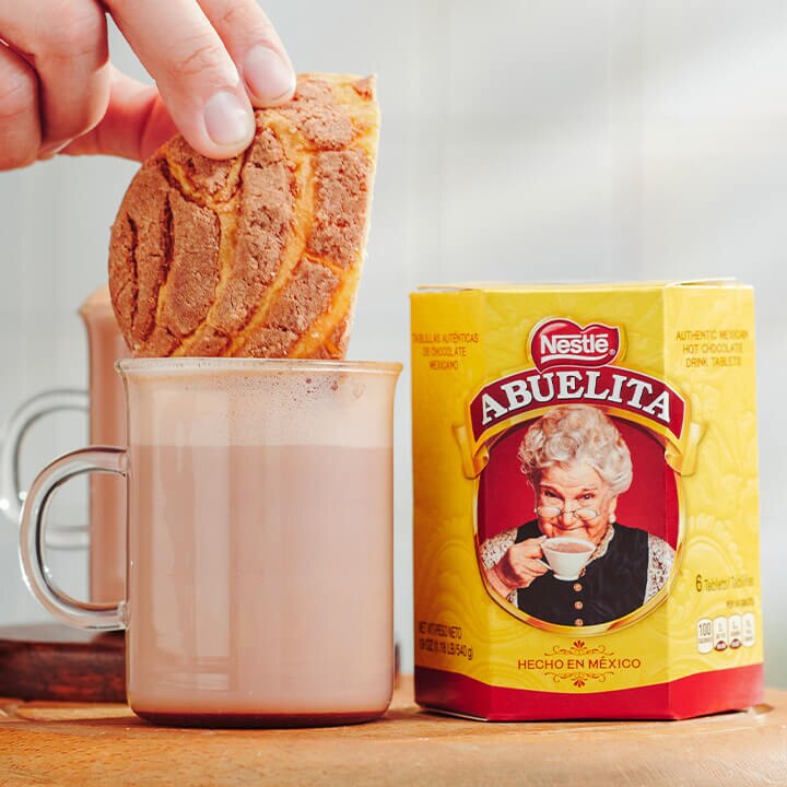 A pitcher pours coconut chocolate milk into glass jars beside a package of ABUELITA tablets.