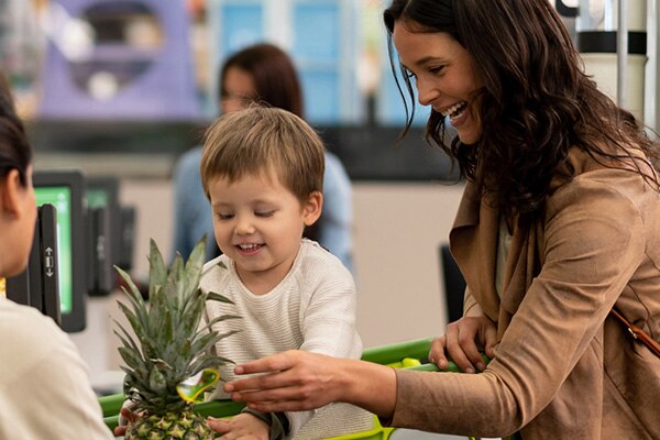 A young boy sitting in a grocery shopping cart helps his smiling mother pass a fresh pineapple to a cashier at the checkout.