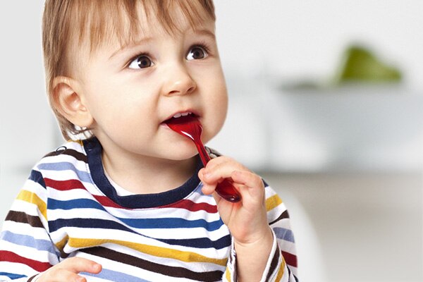 A toddler in a striped shirt chews on a red spoon.