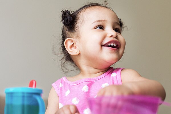 Photograph of a 1 year-old girl smiling in a pink headband, white long sleeve shirt, and pink pants.