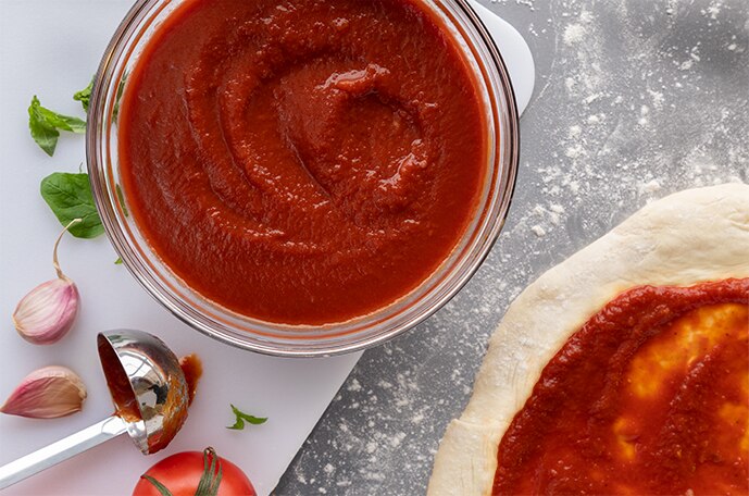 A bowl of pizza sauce surrounded by garlic, herbs, tomatoes and pizza crust with sauce.