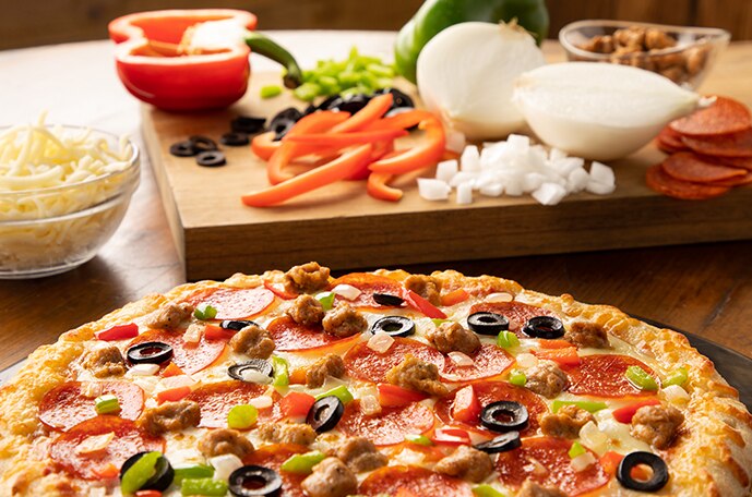 A supreme pizza in front of a cutting board with onions, peppers, sausage, pepperoni and cheese.