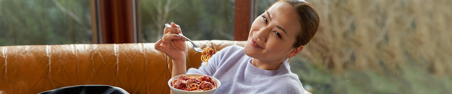 Mujer reclinada sobre un sillón anaranjado sonriendo a la cámara y a punto de probar espagueti y albóndigas Lean Cuisine.