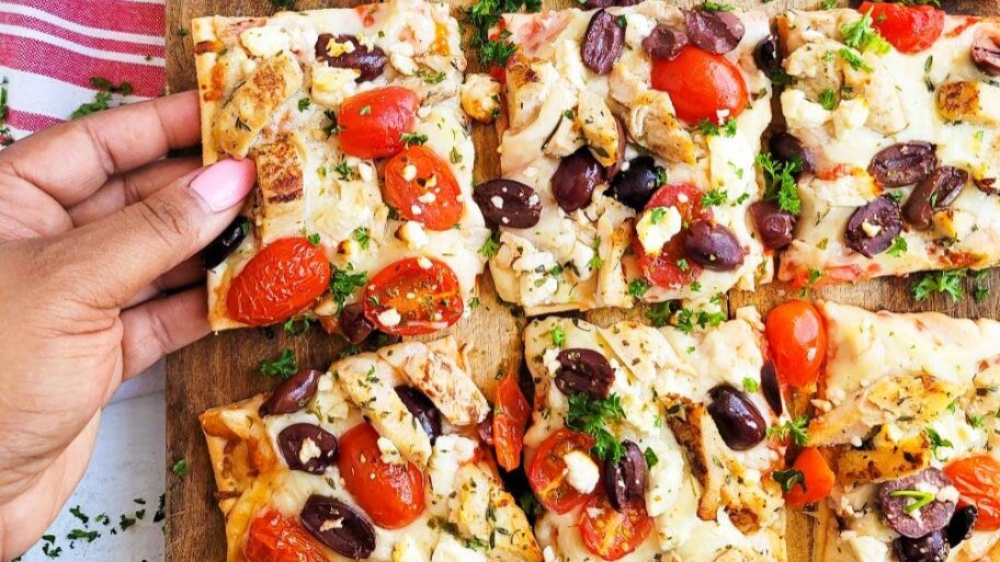A hand reaches for a DiGiorno Thin Crust pizza topped with chicken, feta cheese, kalamata olives and cherry tomatoes.