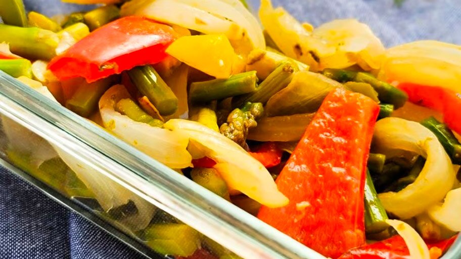 A casserole dish of cooked peppers, onions and asparagus on a cloth napkin near a wooden spatula.