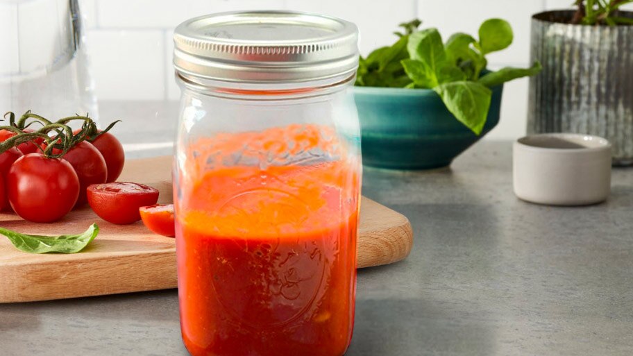 A jar of tomato sauce on a cloth napkin near a wooden spoon and scattered herbs.