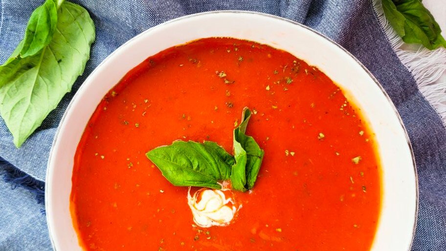 A bowl of creamy tomato bisque with basil garnish surrounded by basil leaves and a cloth napkin.