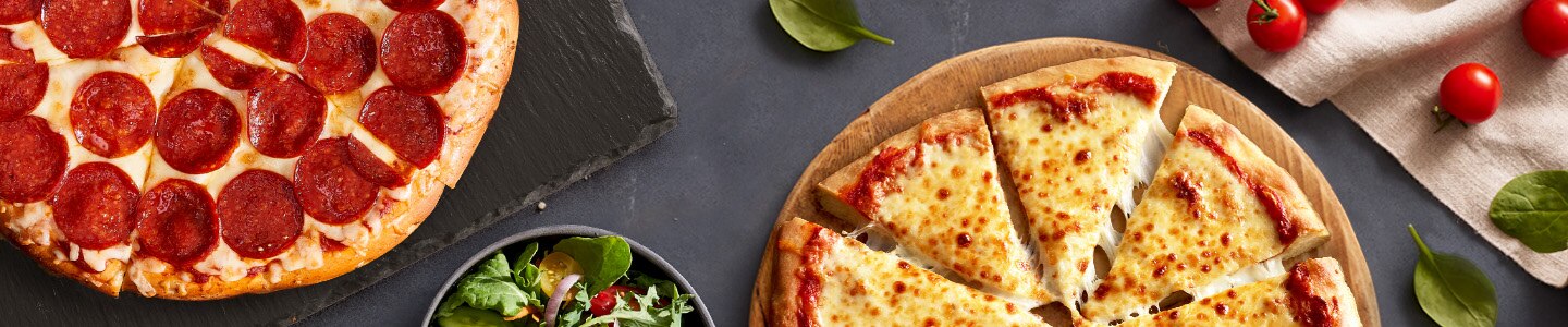 Pepperoni and Cheese Pizzas on cutting boards next to a salad, baby tomatoes, and a basil leaf on a grey background.