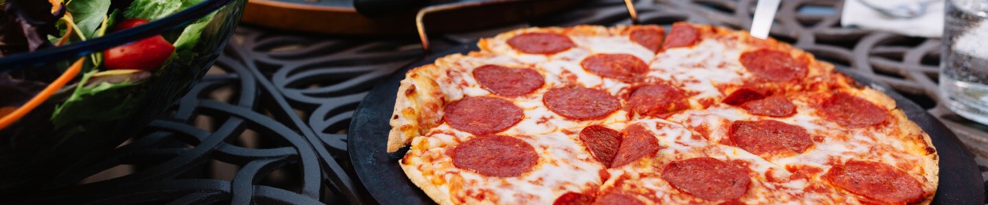 Pizza de pepperoni sobre un plato negro cerca de una ensalada de la casa sobre una mesa con rejillas.