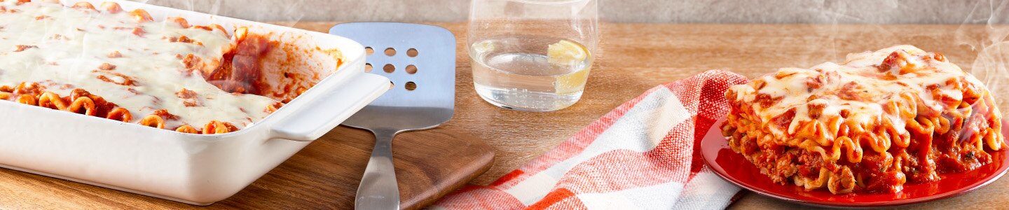 Lasagna in a casserole pan next to a glass of water, spatula, and napkin on a wood table background.