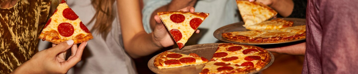 Pizza de pepperoni y queso en bandejas, y al fondo personas tomando rebanadas.